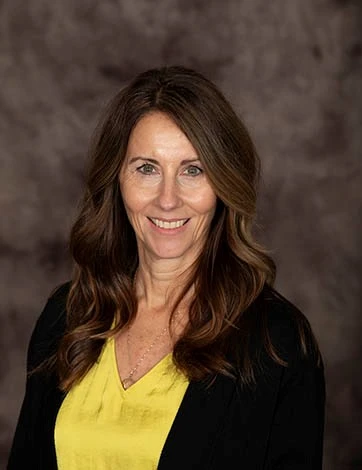 This image is a headshot of a women with grown hair wearing a yellow top and black cardigan. 